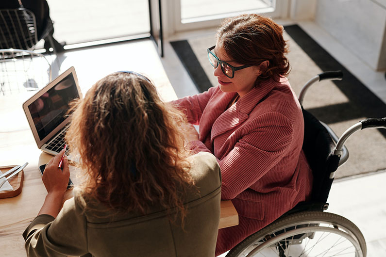 A woman in a wheelchair at a desk speaking to another woman