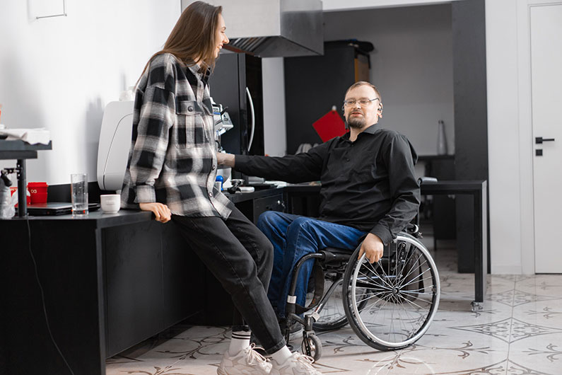 A man in a wheelchair and a woman sitting on his desk speak in an office