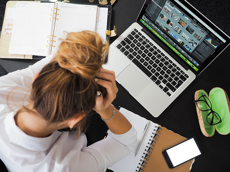 A woman at her desk holding her head in her hands is seen from above